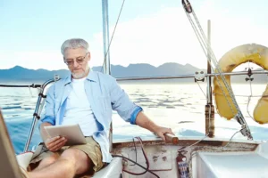 A man wearing sunglasses reads a book or newspaper on a yacht or sailboat on a sunny day.