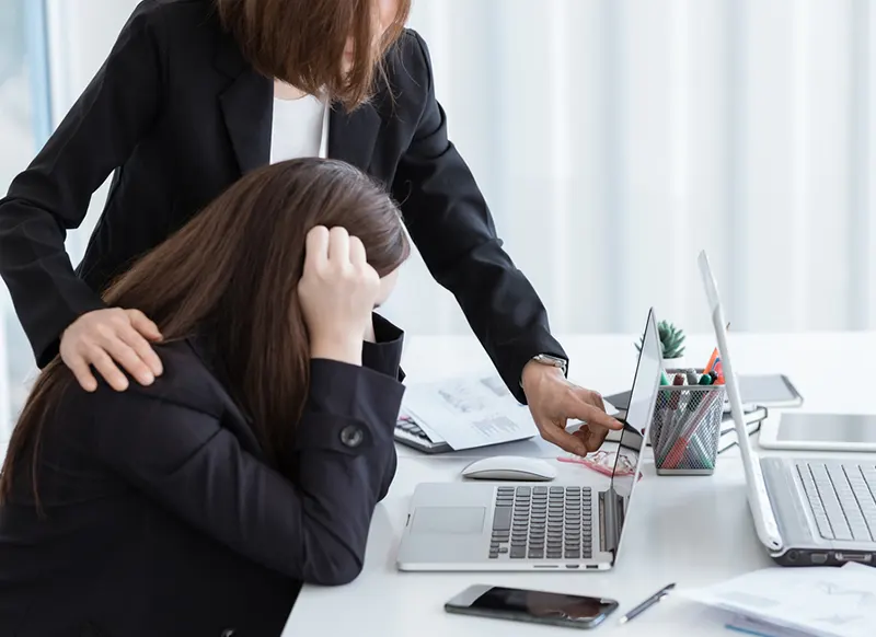 A frustrated knowledge worker clutches her hair while a female colleague stands next to her with a hand on her shoulder and points at something on her laptop screen