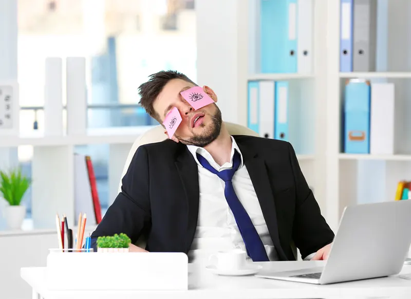 A businessman with a loose tie sleeps sitting down at his desk in front of a laptop has hand-drawn eyes on Post-it notes covering his own eyes