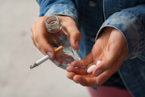 person holds liquor bottle, cigarette, pills