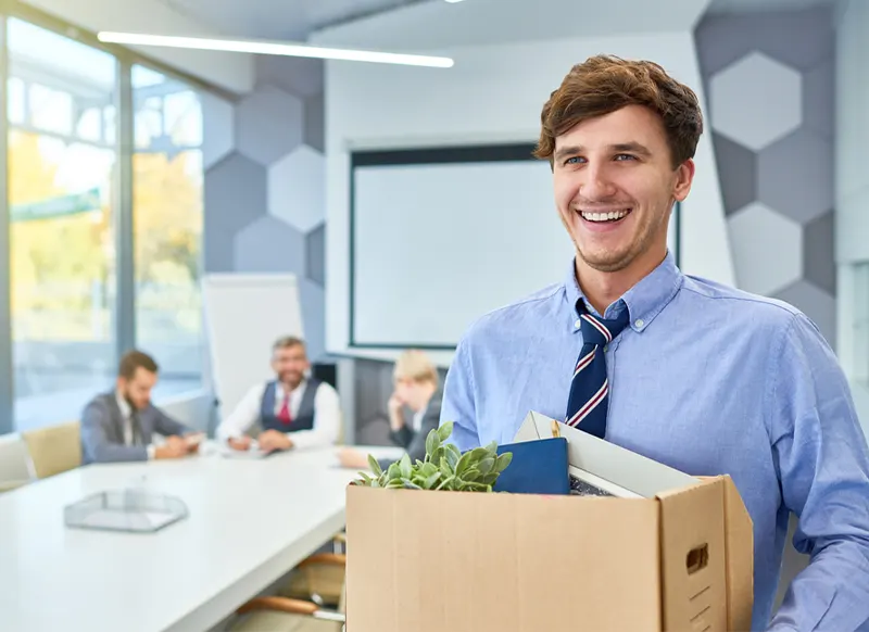A smiling man wearing a long-sleeved blue shirt and a loose striped tie carries a cardboard box of his belongings after being fired or laid off or quitting