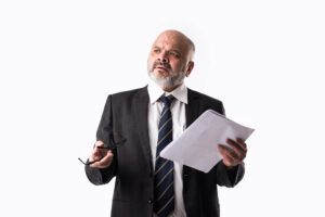 A businessman in a suit and tie holds his glasses and a stack of papers while thinking about what to do and say next