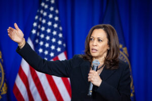 U.S. presidential candidate Kamala Harris speaks into a microphone while holding up one arm in front of an American flag