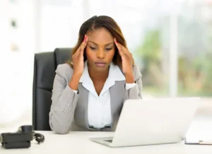 A female person of color sitting in a chair in front of a laptop holds her hands to her temples with a stressed facial expression