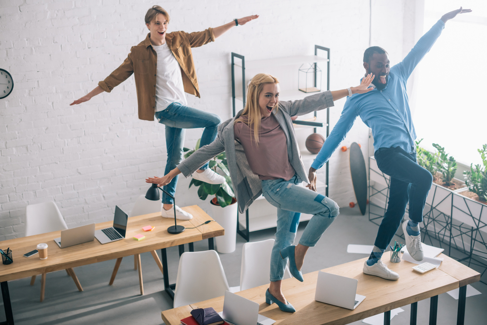 Three happy employees in colorful, casual attire smile broadly and do a coordinated dance on top of their desks