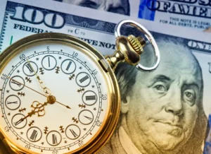 A fancy old-fashioned golden watch with Roman numerals rests on a stack of U.S. $100 bills with Benjamin Franklin's face on them.