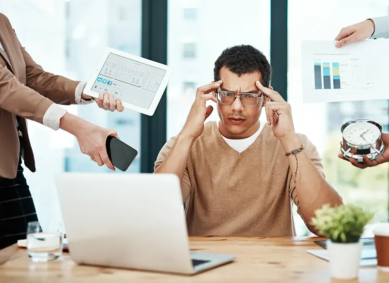 A man with glasses presses his fingers into his forehead and frowns as people on either side of him hold up mobile devices, an alarm clock, and graphs