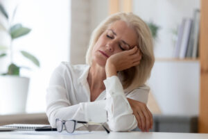 An elderly woman dressed in white who has set down her glasses on a desk closes her eyes and leans her head against one of her hands