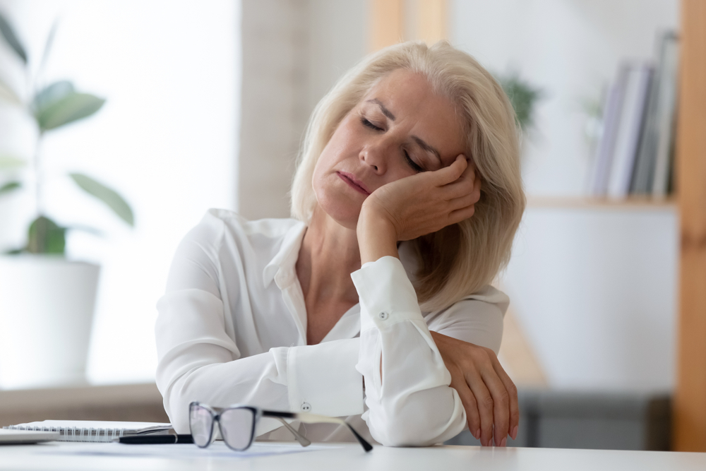 An elderly woman dressed in white who has set down her glasses on a desk closes her eyes and leans her head against one of her hands