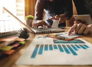 Two employees collaborate, one using a pencil and laptop, the other using a calculator and charts