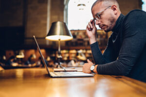 A male professor wearing glasses and a watch looks at a laptop in front of a lamp
