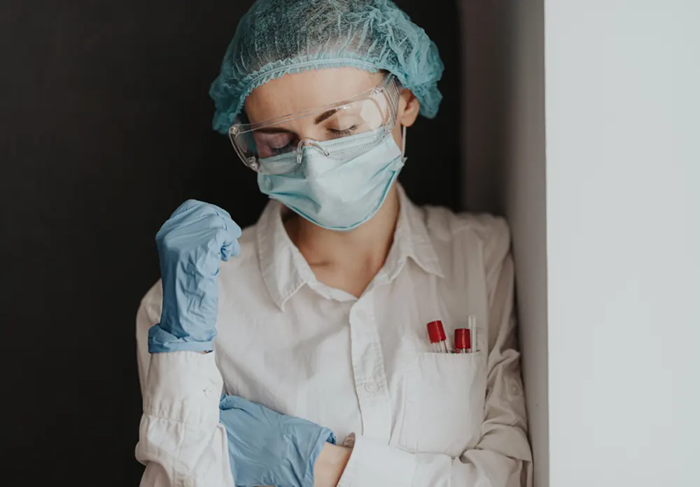 A nurse with closed eyes wears protective gloves, a mask, goggles, and a hairnet