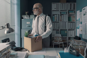 An elderly man with glasses stares through his office's window blinds holding a box of his personal possessions