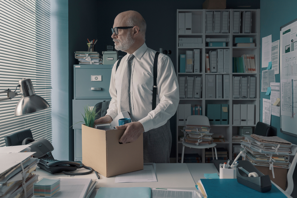 An elderly man with glasses stares through his office's window blinds holding a box of his personal possessions