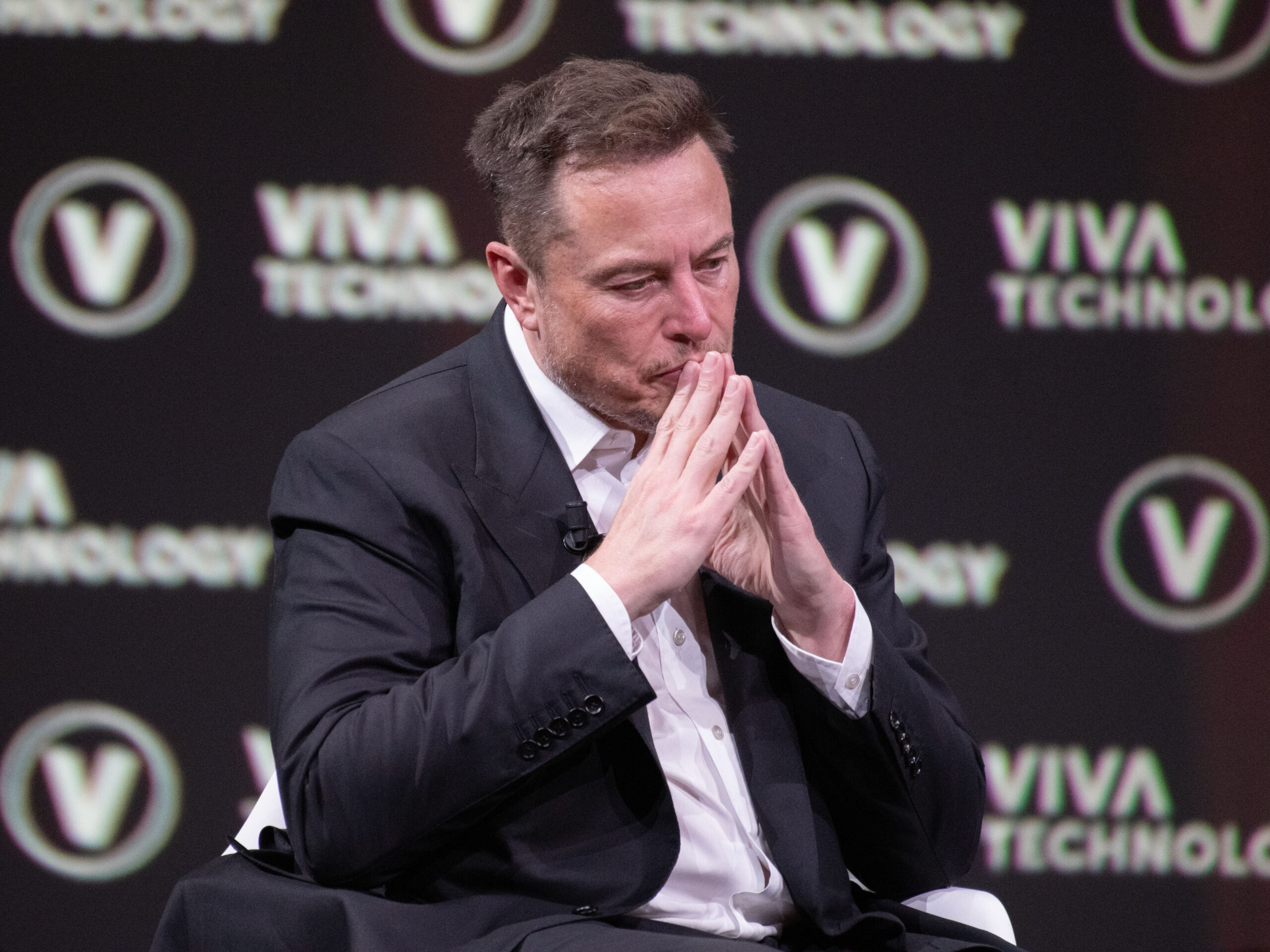Elon Musk, CEO of SpaceX, X.com, and Tesla, thinks while sitting on a stage with his hands pressed together against his lips