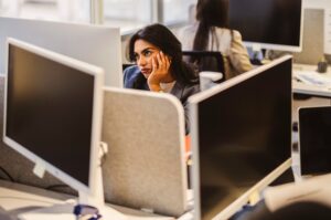 An annoyed businesswoman rests her chin on her hand while sitting at her desk in an office cubicle, lost in thought.