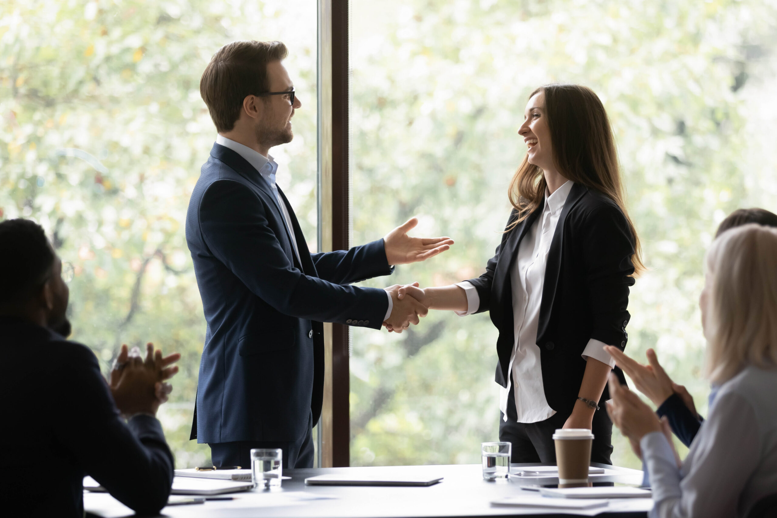 A male leader in glasses and a suit without a tie shakes hands with a young smiling female employee as colleagues applaud