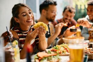 A smiling young woman raises a taco toward her mouth, with drinks and more tacos on the table in front of her, and friends in the background eat chips and salsa
