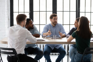 A light-skinned man with glasses and a beard speaks to a diverse group sitting at a table