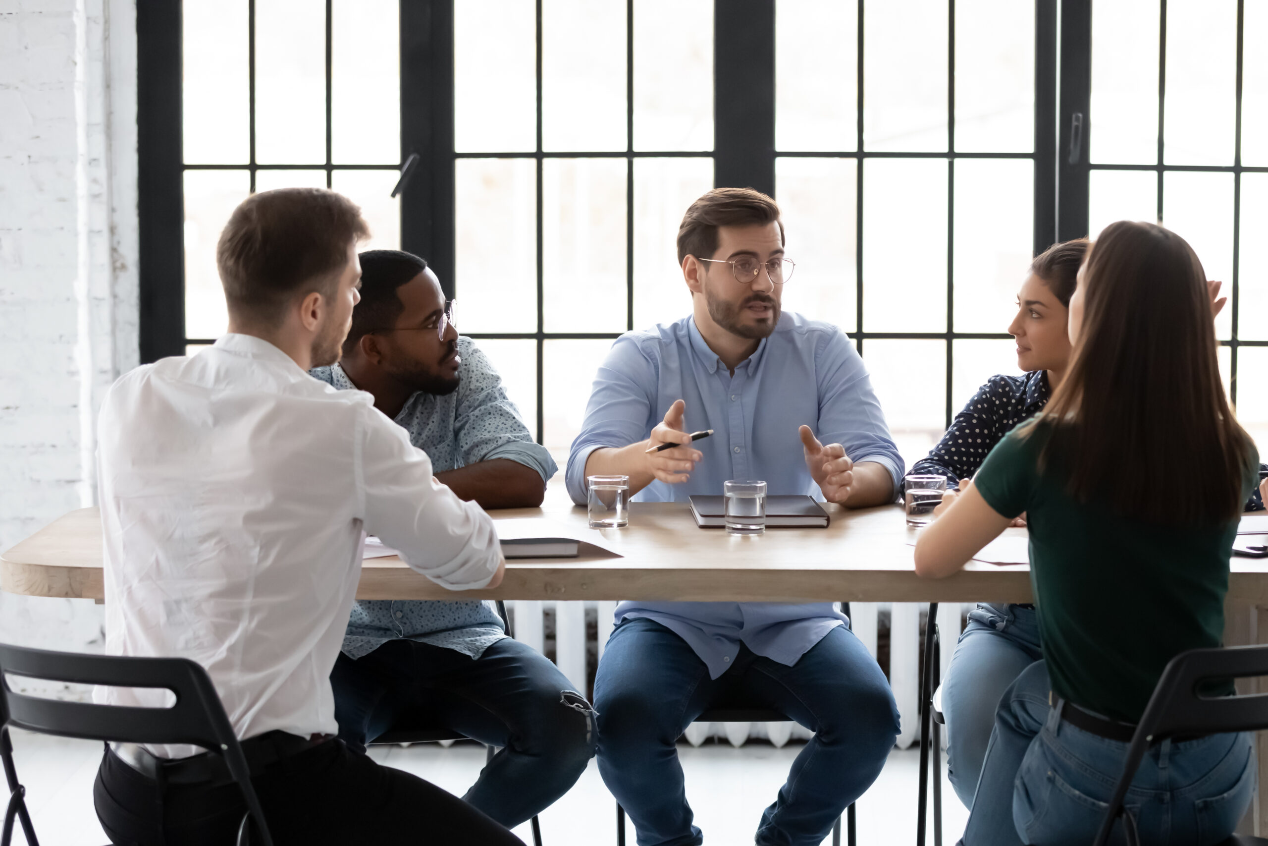 A light-skinned man with glasses and a beard speaks to a diverse group sitting at a table