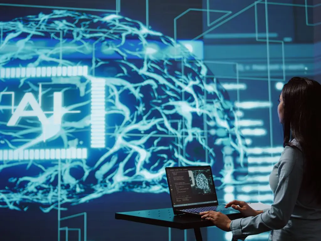 A woman works on a laptop in front of a large screen with a virtual brain labeled AI