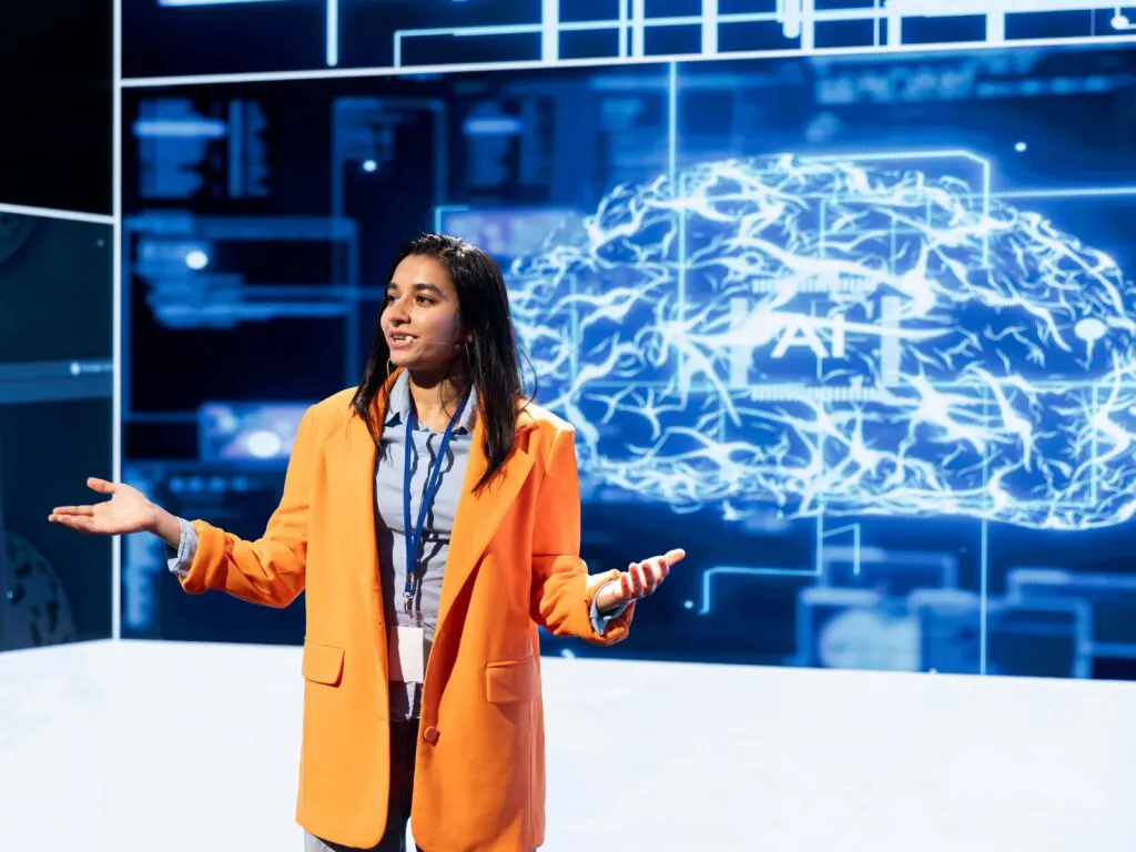 A woman in an orange jacket wearing a conference lanyard and name badge talks about AI tools