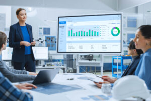 A female executive in a blue suit presents data, including charts and graphs, on a whiteboard in a meeting room with male and female colleagues.