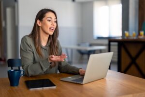 A woman working from home speaks to someone in a virtual meeting on her laptop, gesturing with her hand