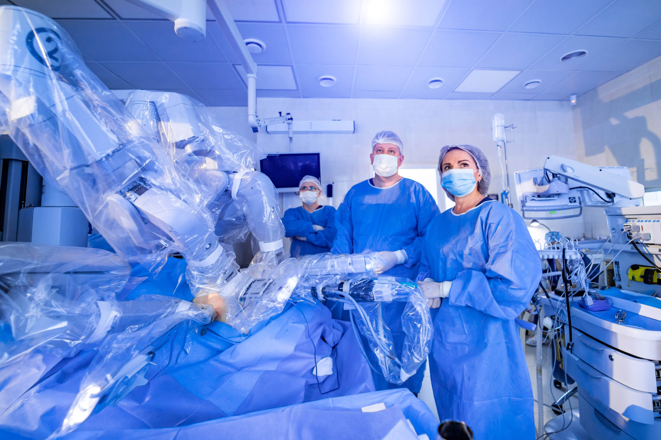 Three surgeons in operating-room scrubs and masks monitor a robotic surgical system in a hospital