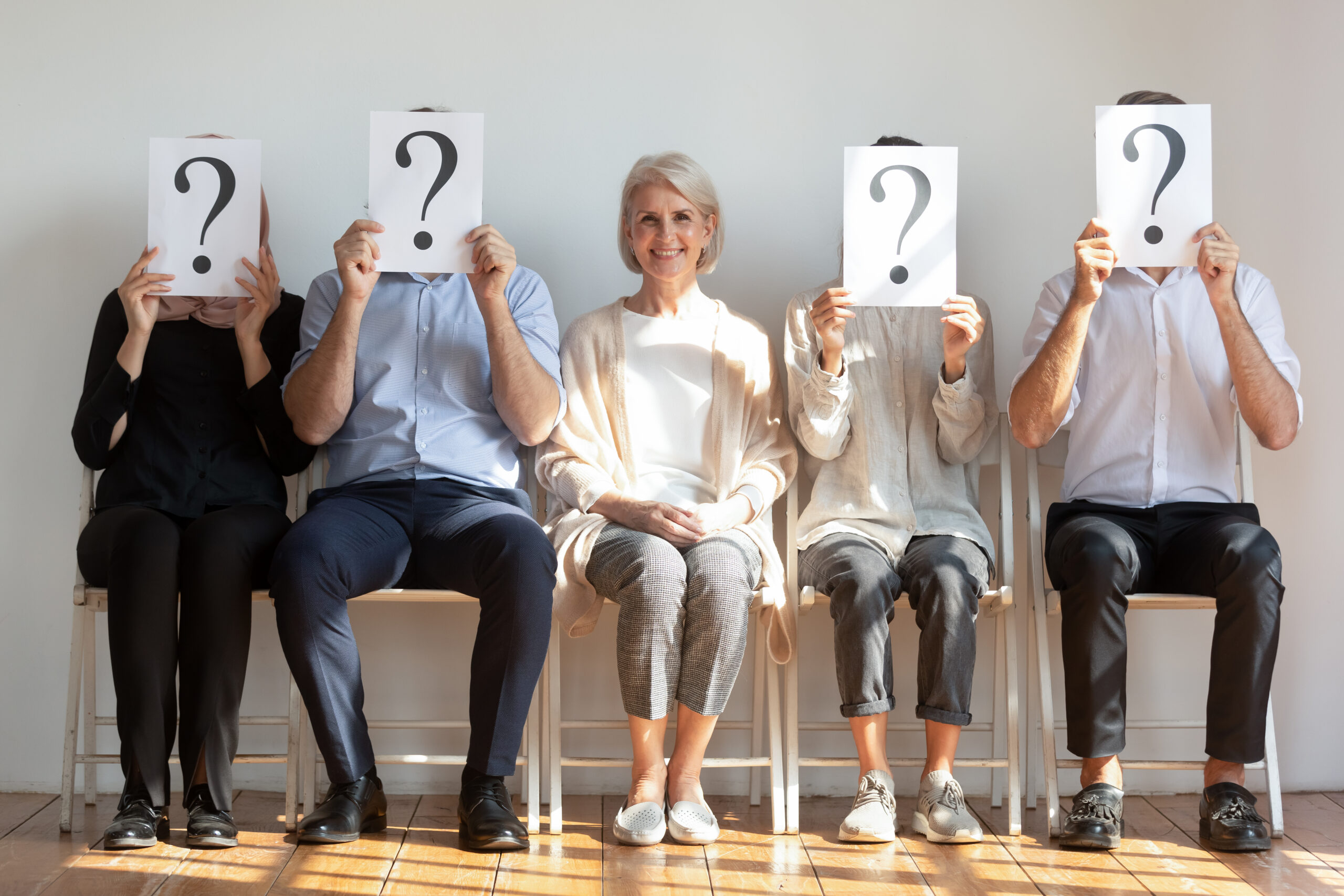A female candidate waits to be called in for a job interview while candidates holding a question mark in front of their faces sit next to her.