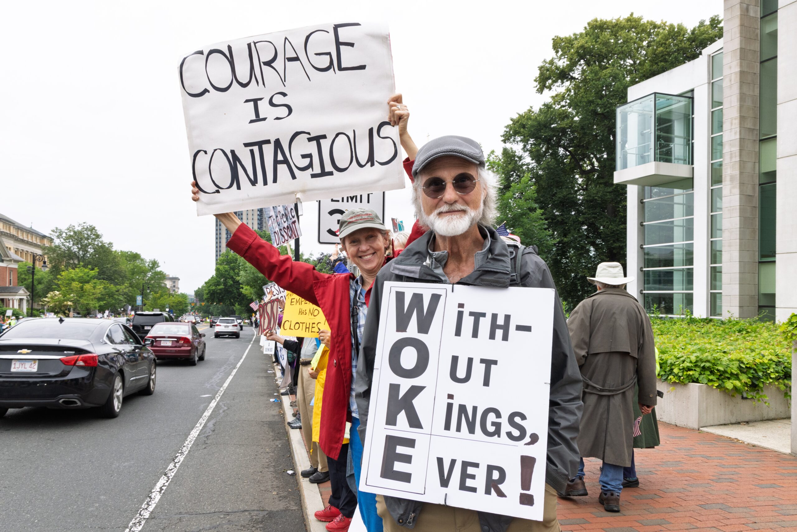 People at a political protest hold signs that read "Courage Is Contagious" and "WOKE: With- Out Kings Ever!"