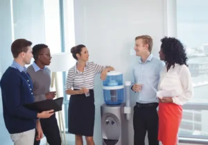 A diverse group of professionally dressed colleagues talks around the watercooler in an office.