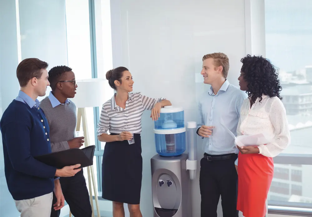 A diverse group of professionally dressed colleagues talks around the watercooler in an office.