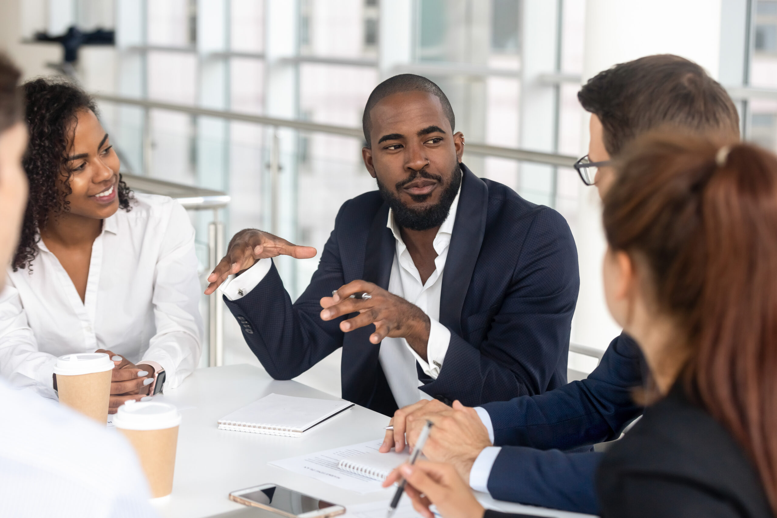 An African American entrepreneur or CEO in a suit without a tie talks about business insights surrounded by men and women listening attentively.
