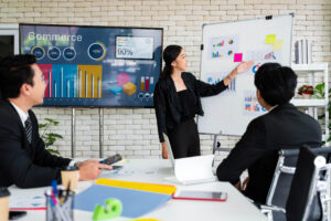 A cheerful and confident Asian businesswoman stands, present bar charts data from projector screen to her office colleagues. Asian business women leader role at the meeting.