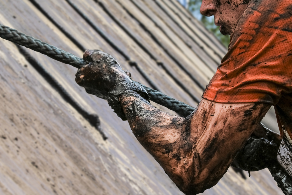 A dirty wiry muscular man competes in a mud race, climbing a wall by pulling on a rope.