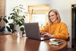 A happy mature woman in an orange sweater uses a laptop while working remotely from home next to a green leafy plant.