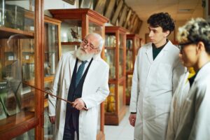 Senior Caucasian man explaining scientific exhibit to two teenage boys, both wearing lab coats, standing in museum display room with glass cabinets