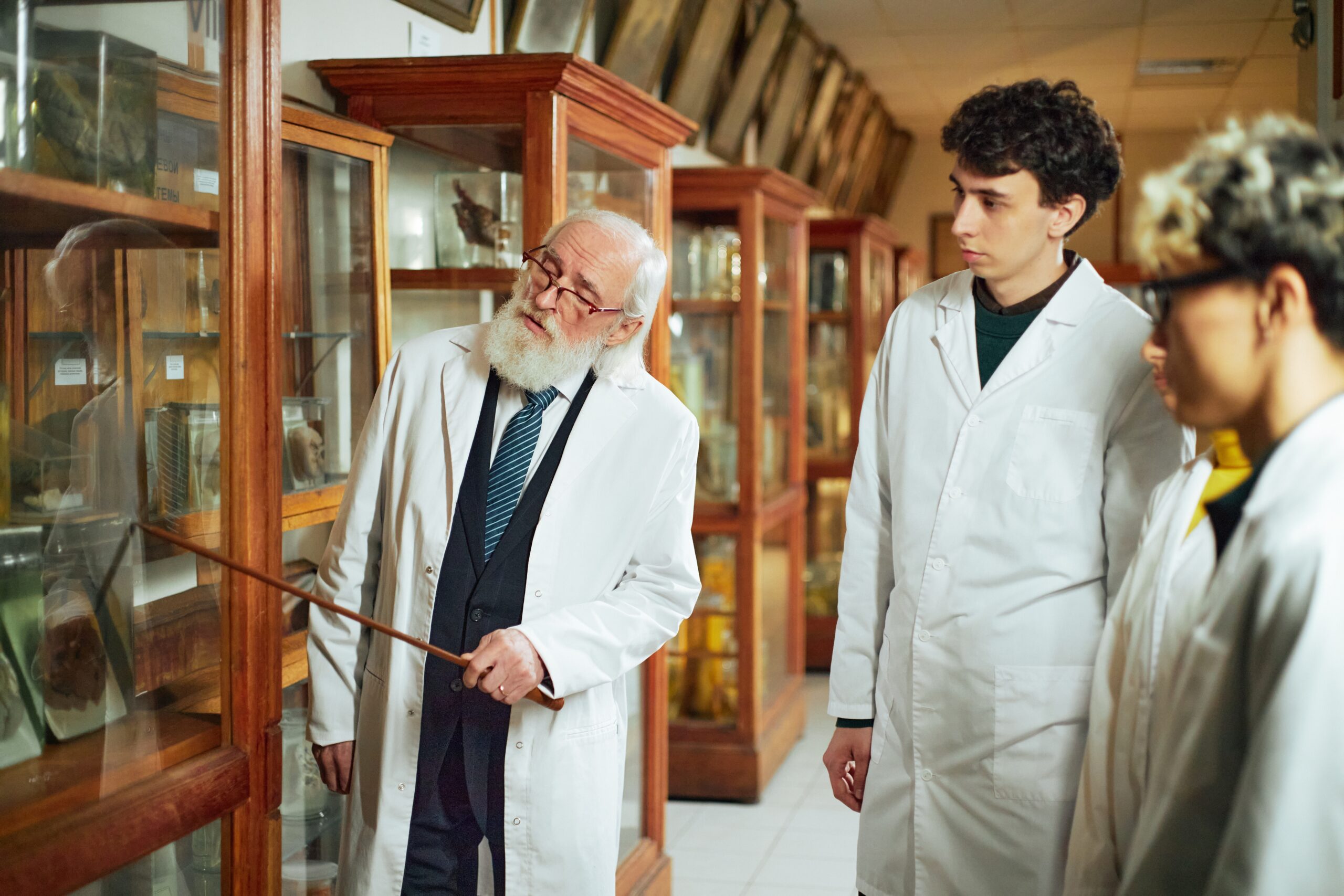 Senior Caucasian man explaining scientific exhibit to two teenage boys, both wearing lab coats, standing in museum display room with glass cabinets