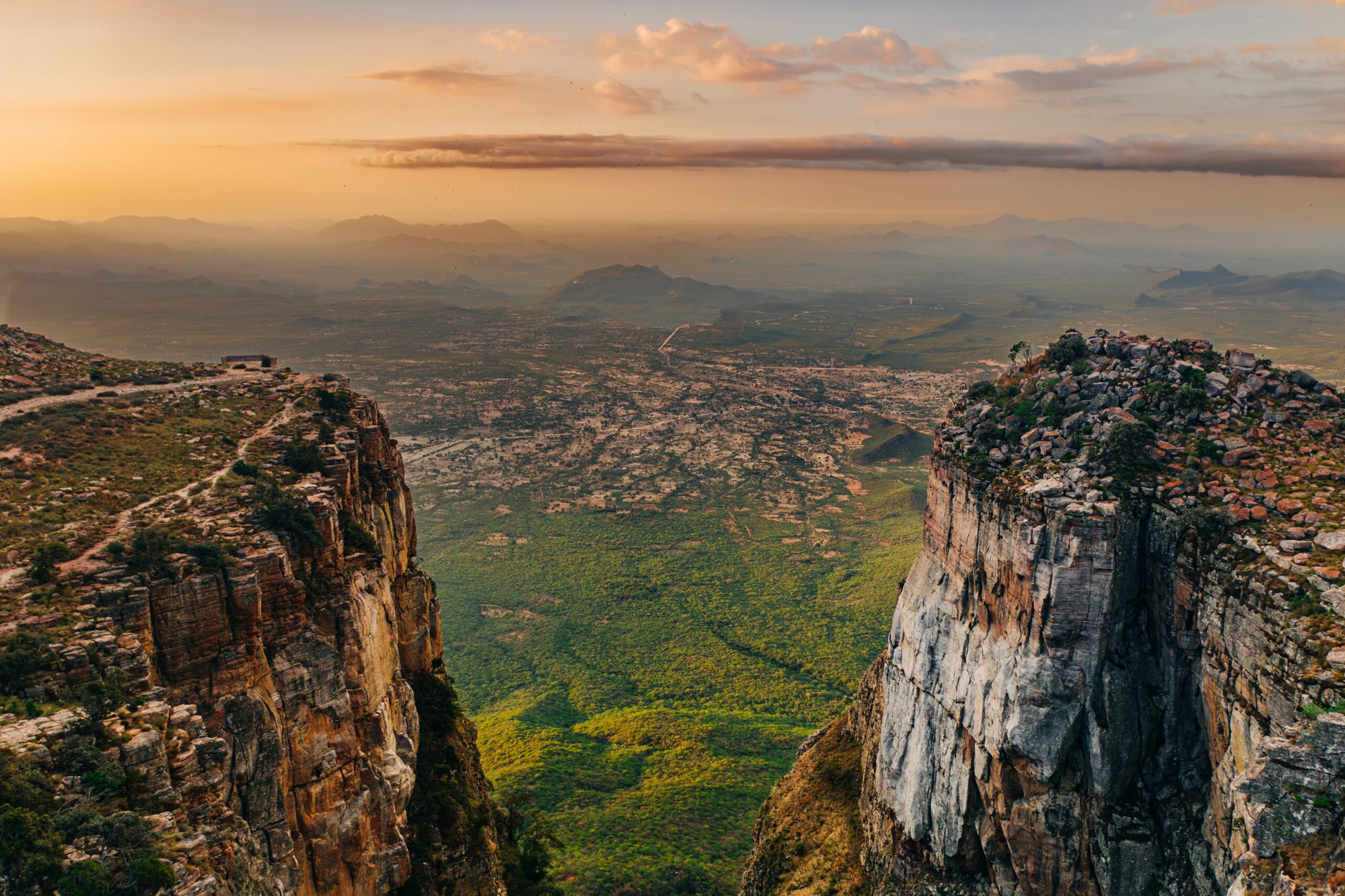 Aerial view from the top of Serra da Leba escarpment overlooking the vast plains and dramatic cliffs of the Tundavala Gap, with distant mountains under warm sunset light in Huíla Province, Angola.