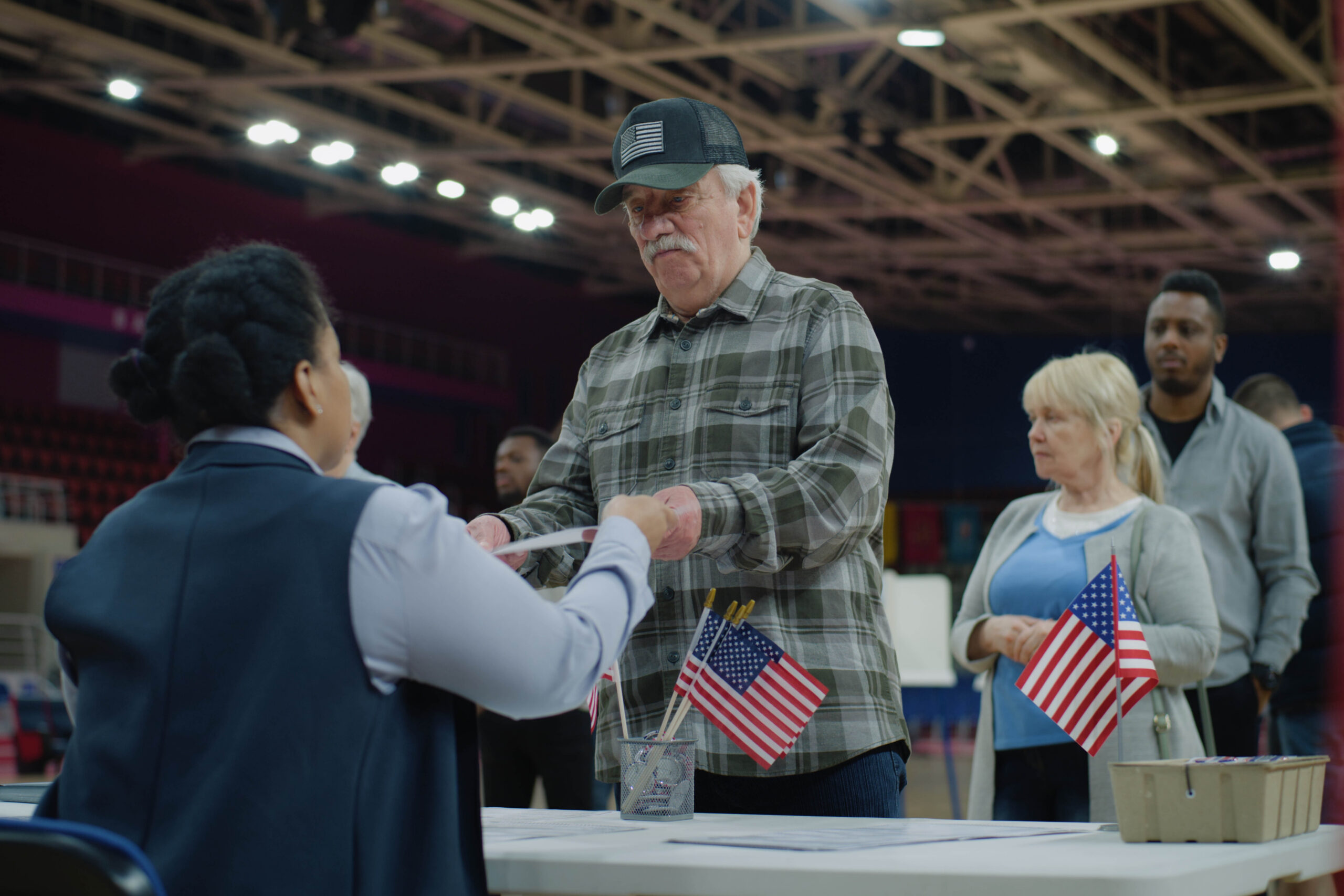 Elderly man talks with polling officer and takes vote bulletin. Multi cultural American citizens come to vote in polling station. Political races of US presidential candidates. National Election Day