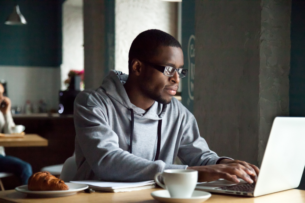 Black man dressed in business casual gray shirt and jacket looking at a laptop screen.